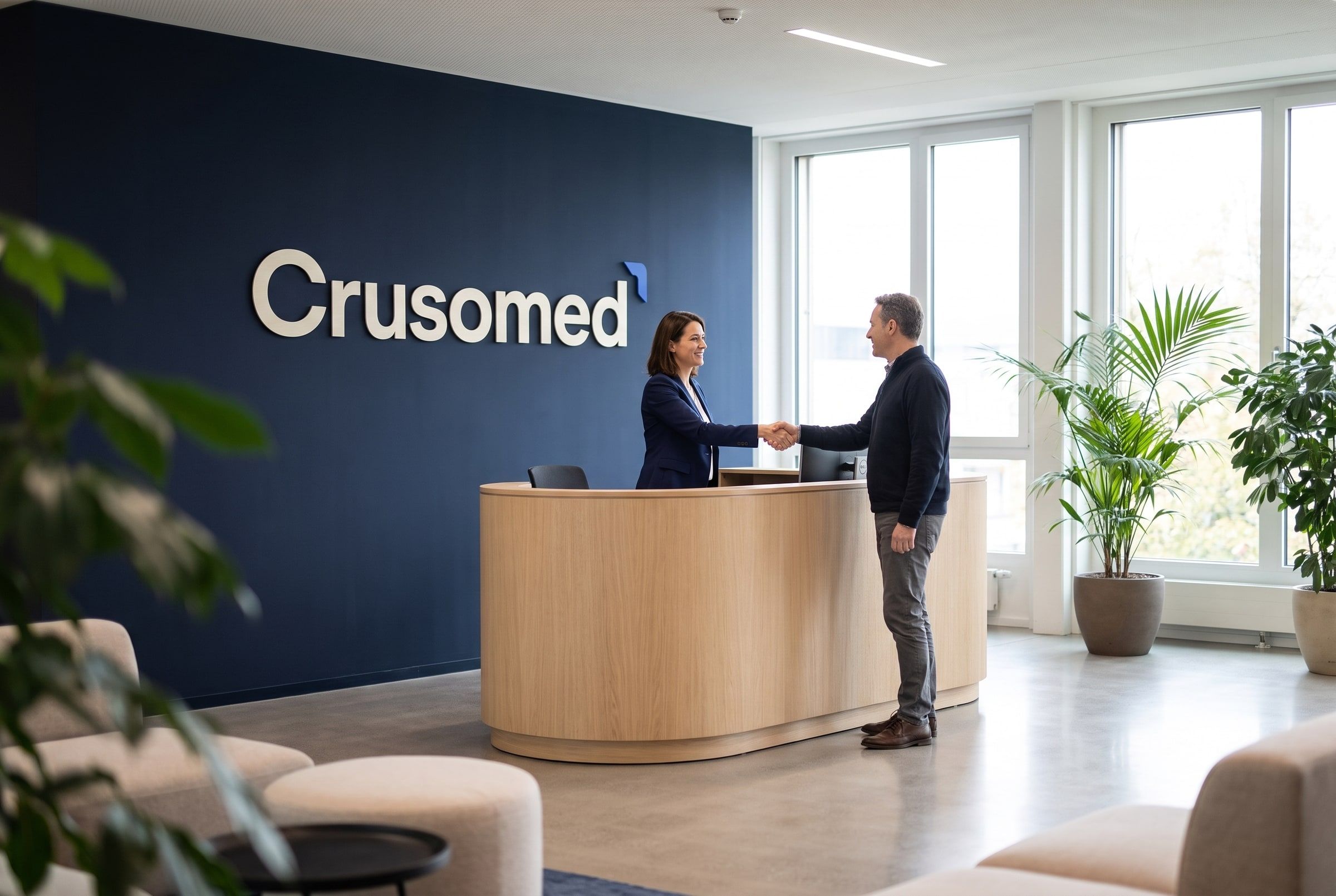 Two professionals greeting each other in a bright modern office reception, the Crusomed wordmark on the deep navy accent wall behind the desk.