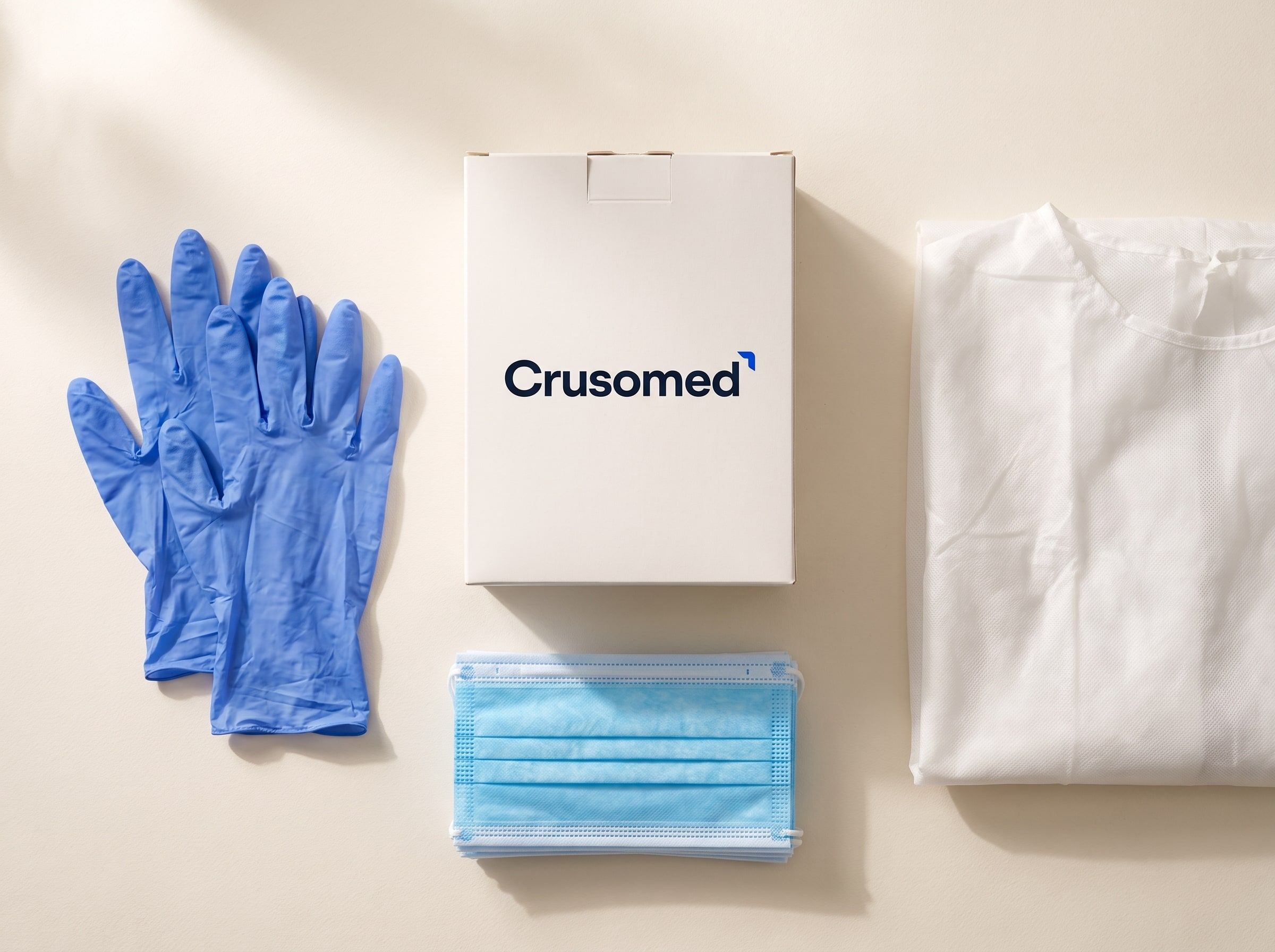 Protective equipment arranged on a light surface: examination gloves, face masks, a folded gown and a Crusomed-branded dispenser box.
